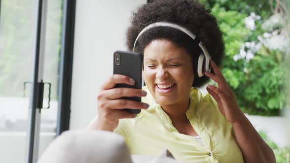 Video of happy plus size african american woman with headphones on sofa, listening to music