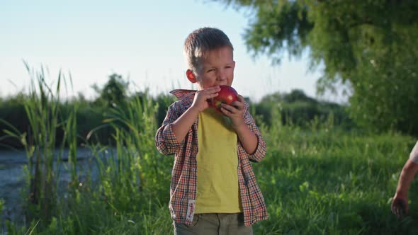 Little Boy Happily Eating a Juicy Ripe Apple While Relaxing Near the River Background of Trees and