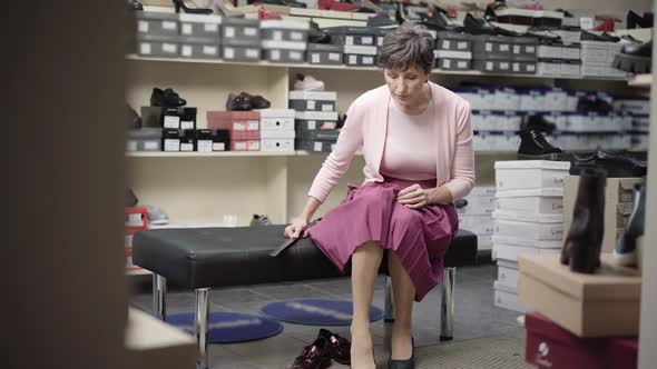 Wide Shot of Serious Caucasian Senior Woman Sitting in Shoe Store and Trying on High-heels. Portrait