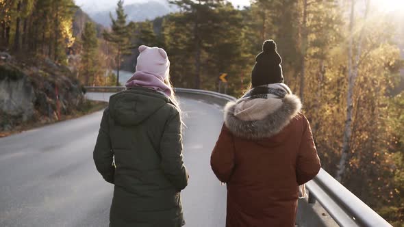 Two Female Tourists Walking in Slow Motion on a Long Road Through the Countryside Towards the Snowy