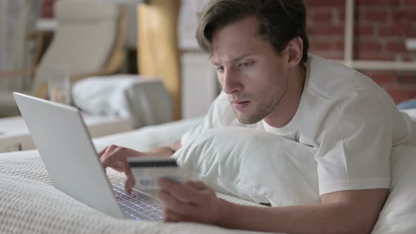 Successful Young Man Using Credit Card on Laptop in Bed
