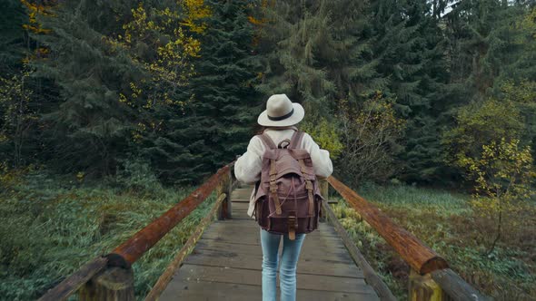 Back View Camera Follows Hipster Woman in Casual Wear and Hat with Backpack Hiking in Forest in