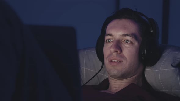 Smiling Young Man in Headphones Watching Film Movie on Laptop While Resting on Bed at Night