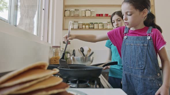 Two young girls preparing pancakes in the kitchen using a frying pan