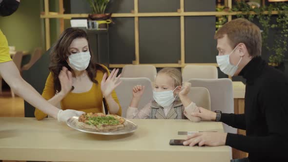 Positive Family in Covid Face Masks Sitting in Restaurant Unrecognizable Waiter Bringing Fresh Baked