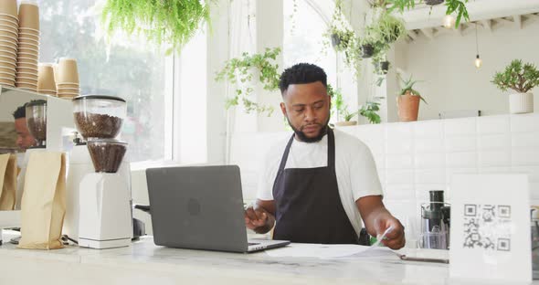 African american male cafe owner using laptop and looking at paperwork behind counter at cafe