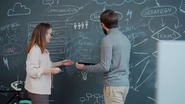 Girl and Guy Discussing Business Strategy and Writing on Chalkboard Wall Indoors