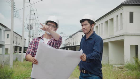 Two Asain confident engineers discussing blueprint while standing at construction site