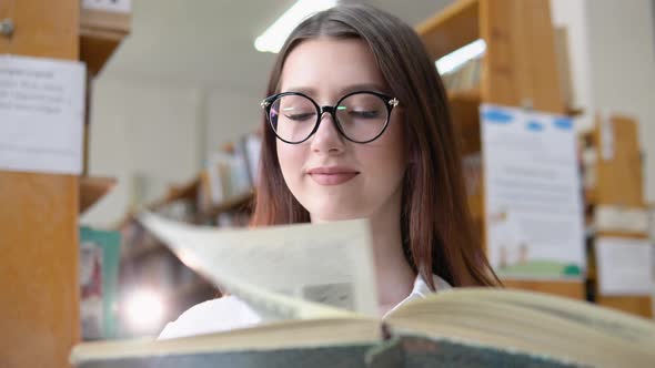 A Smiling Young Girl in Glasses Reads a Book in the University Library
