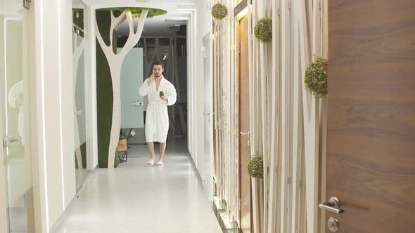 Young Man in White Robe Walks Down the Hall of Luxurious Spa Center Talking on Mobile Phone.