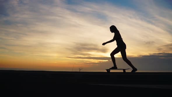 Silhouette of a Girl in Shorts and Sneakers Skateboarding Along the Road Against the Ocean and