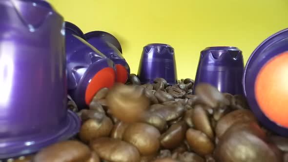 Dolly shot of falling roasted coffee beans in slow motion surrounded by coffee capsule. Yellow backg