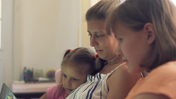 Two Girls And Mother Play On Tablet At Home On Sofa