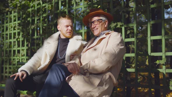 Senior Father and Young Hipster Son Sitting on Bench in Park and Talking