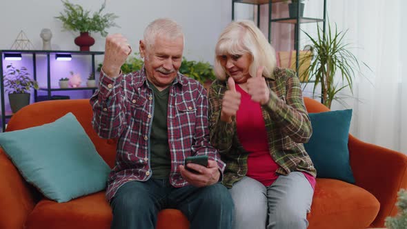 Overjoyed Grandmother Grandfather Hold Smartphone Excited About Mobile App Sport Bet Bid Win at Home