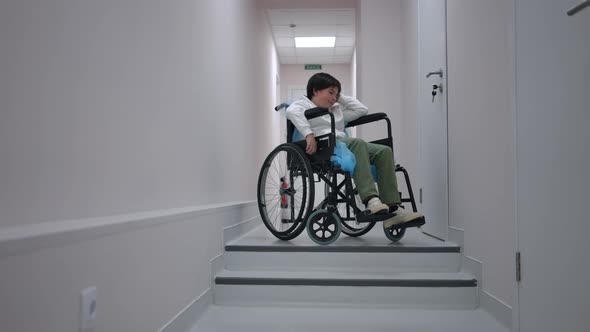 Wide Shot Portrait of Sad Thoughtful Caucasian Boy in Wheelchair Sitting at Stairs in Hospital