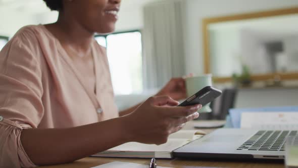 Happy african american woman sitting at table using smartphone and drinking coffee