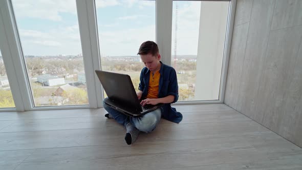 Teen boy sitting on the floor by the window and playing on laptop. 