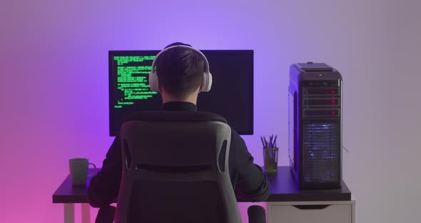 Rear View of Man Programmer in Headphones Coding Network Security Software Indoors in Neon Lights at