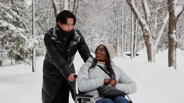 Young Couple of Asian Man and His Black Girlfriend in the Wheelchair Talking on the Snowy Path