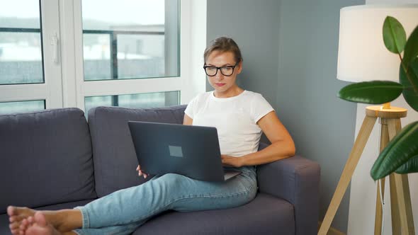 Woman with Glasses is Sitting on the Couch and Working on a Laptop