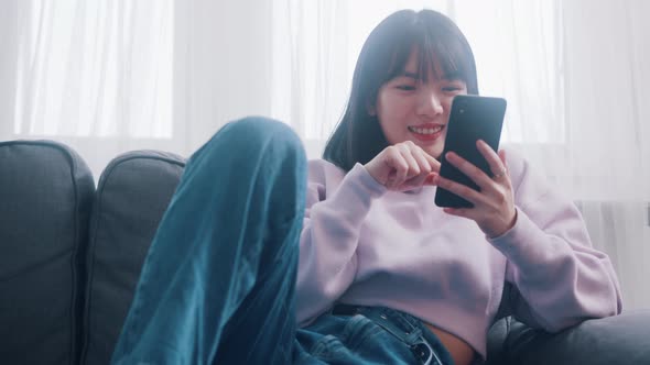 Young Happy Asian Woman Relaxing on the Couch and Browsing Social Media on Her Smartphone