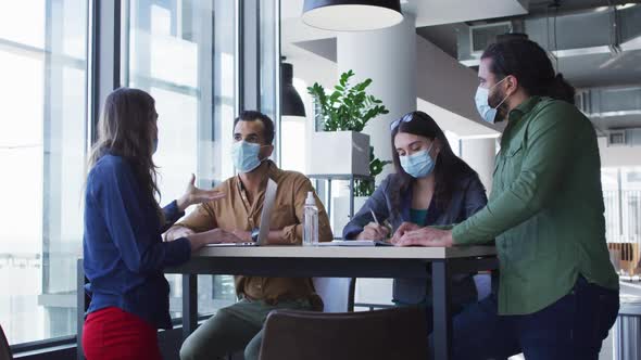 Diverse group of business colleagues wearing face masks sitting at table and talking