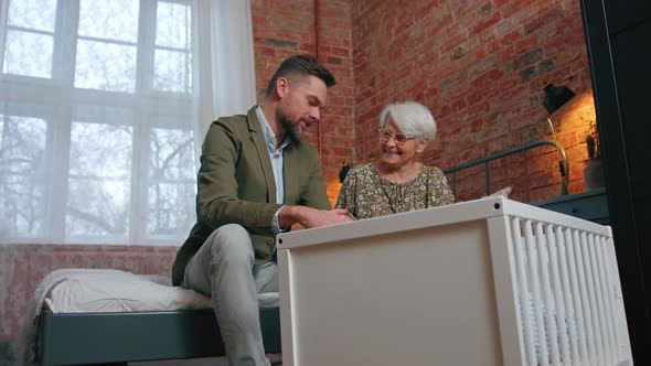 Elderly Caucasian Pensioner Grandma and Her Middleaged Bearded Son Looking Cheerfully at Newborn