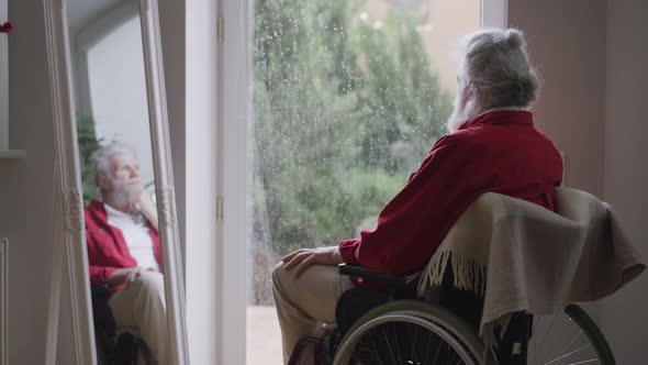 Sad Disabled Bearded Greyhaired Old Man Looking at Rain Sitting in Wheelchair Indoors