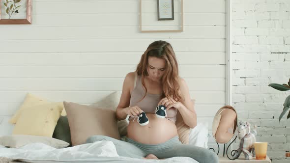 Closeup Expecting Mother Playing Baby Shoes on Pregnant Belly in Bed.