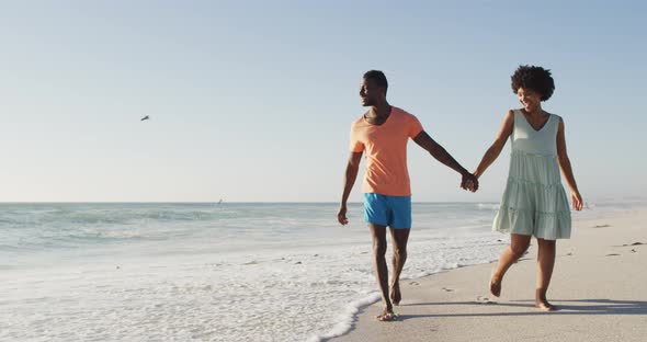 Smiling african american couple holding hands and walking on sunny beach