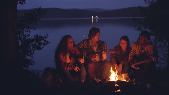 Happy and Relaxed Guys and Girls Are Singing While Their Friend Is Playing the Guitar Near Campfire