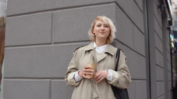 Young Blonde Business Woman in a Beige Trench Coat and White Blouse with a Black Bag and Takeaway