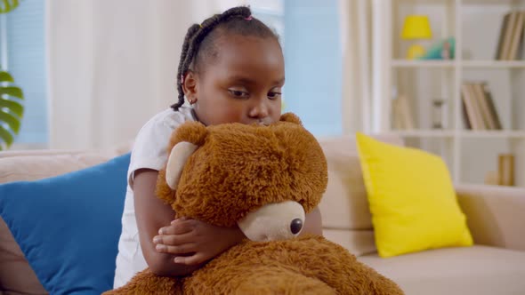 Unhappy Black Kid Girl Hugging Teddy Bear Sitting on Couch at Home Alone