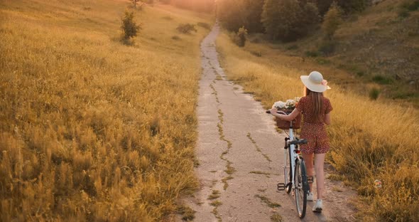 Back Plan Beautiful Blonde in Dress and Retro Bike Walking on the Road in the Summer Field