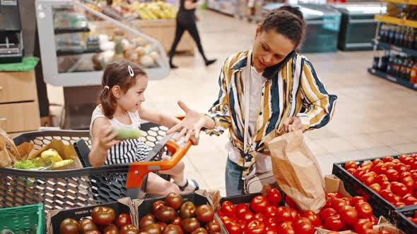 Little Girl with Her Mother in the Supermarket Sitting in a Cart and Helping Her Mother to Choose