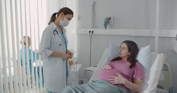 Doctor Wearing Protective Mask Consulting Pregnant Woman Resting in Hospital Bed