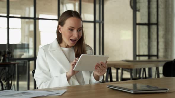 Young Woman Happily Enjoys Winning Using a Tablet While Sitting at a Desk in the Office