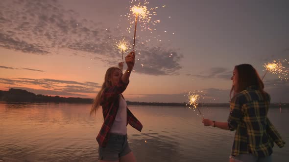 A Group of Friends Girls and Men Dance on the Beach with Sparklers in Slow Motion at Sunset