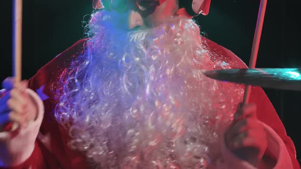 Santa Claus in a Red Costume with Beard and Funny Glasses Plays the Drums in a Dark Studio with