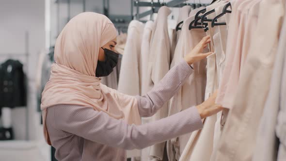 Young Concentrated Arab Woman Shopper in Medical Mask Wearing Hijab Chooses Outerwear in Clothing