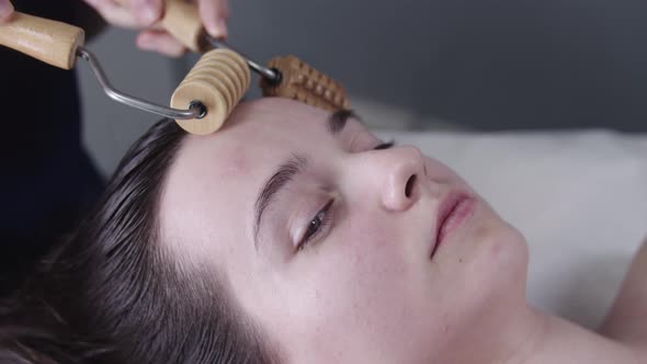 Woman Massagist Using Two Wooden Ribbed Rollers on the Face of Her Female Client