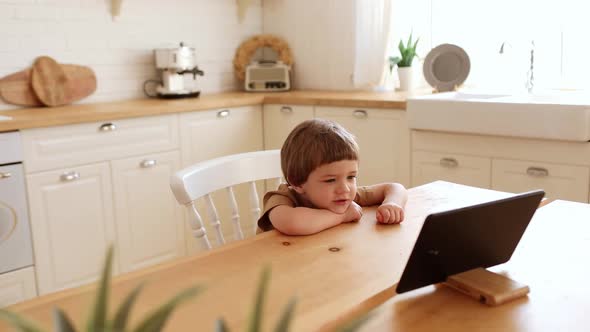 a Little Caucasian Boy Eats Krunchy and Watches Tablet PC at Kitchen Table