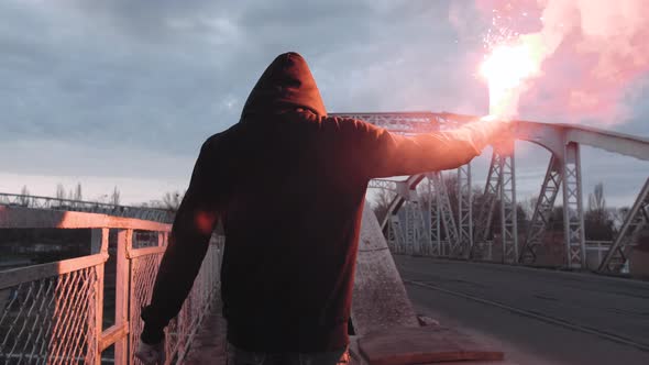 Back View of a Young Man in Hoodie and Balaclava with Red Burning Signal Flare on the Road Under an