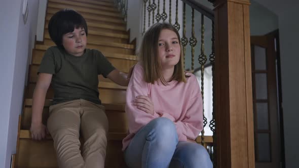 Irritated Teenage Girl Pushing Away Hand of Little Boy Leaving As Sibling Sitting on Stairs Hugging