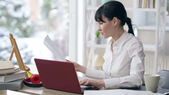 Side View of Confident Focused Young Businesswoman Looking at Documents and Typing on Laptop