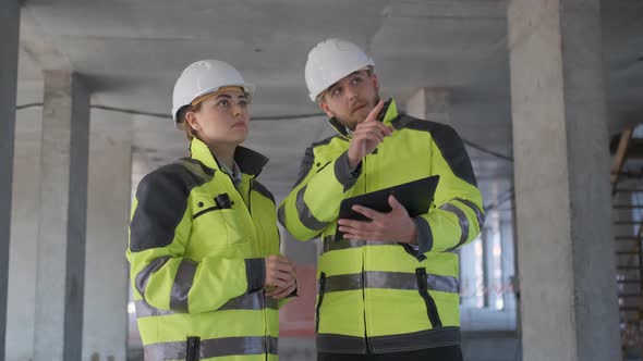 Two Engineer In Helmets With Tablet Device And Big Paper Drawings Discussing