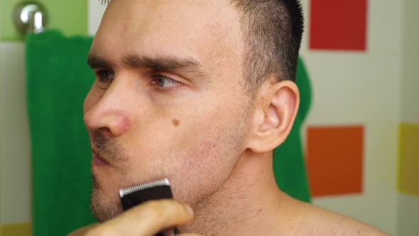 A Caucasian Man Shaves with an Electric Razor Trimmer at Home in the Bathroom