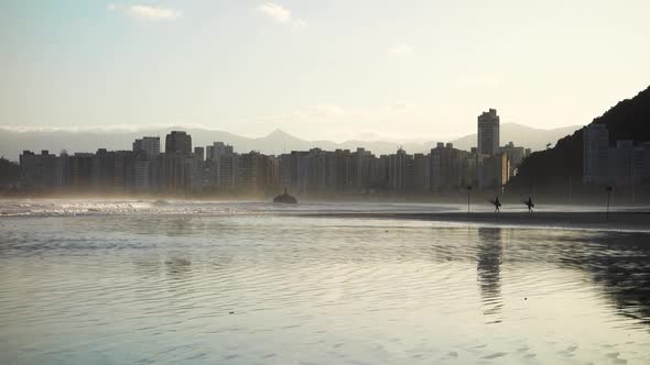 surfers walk on the sand towards sea at dusk. Itarare beach in Sao Vicente. cityscape on background,