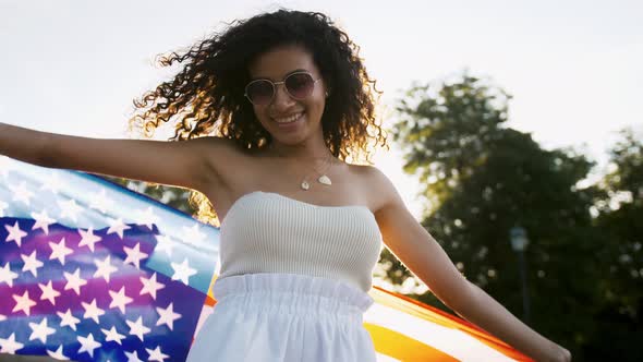 Young Curly Africanamerican Girl in White Outfit and Sunglasses is Holding Flag of USA Smiling and